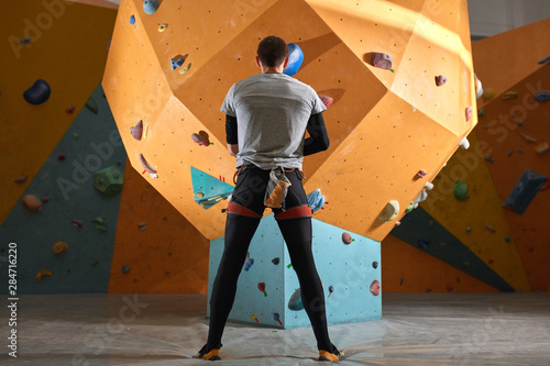Full length shot of physically challenged boulderer standing in front of artificial climbing wall, thinking and planning strategy of climbing up. Low angle back view.