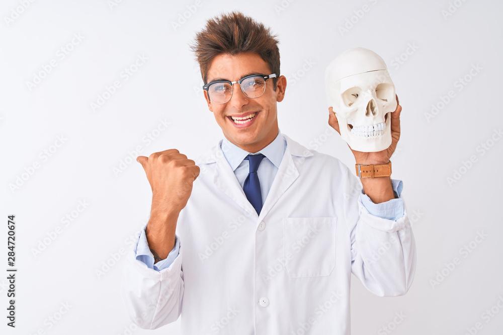 Young handsome doctor man wearing glasses holding skull over isolated white background pointing and showing with thumb up to the side with happy face smiling Young handsome doctor man wearing glasses holding skull over isolated white background pointing and showing with thumb up to the side with happy face smiling