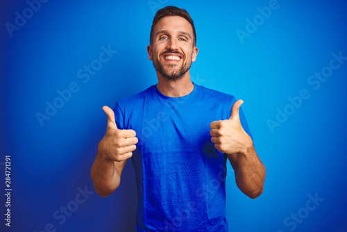 Young handsome man wearing casual t-shirt over blue isolated background success sign doing positive gesture with hand, thumbs up smiling and happy. Cheerful expression and winner gesture.