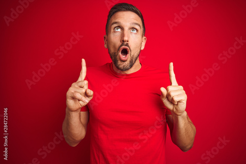 Young handsome man wearing casual t-shirt over red isolated background amazed and surprised looking up and pointing with fingers and raised arms.