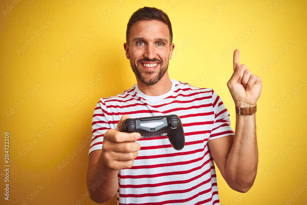 Young handsome man playing video games using joystick game pad over yellow background very happy pointing with hand and finger to the side Young handsome man playing video games using joystick game pad over yellow background very happy pointing with hand and finger to the side