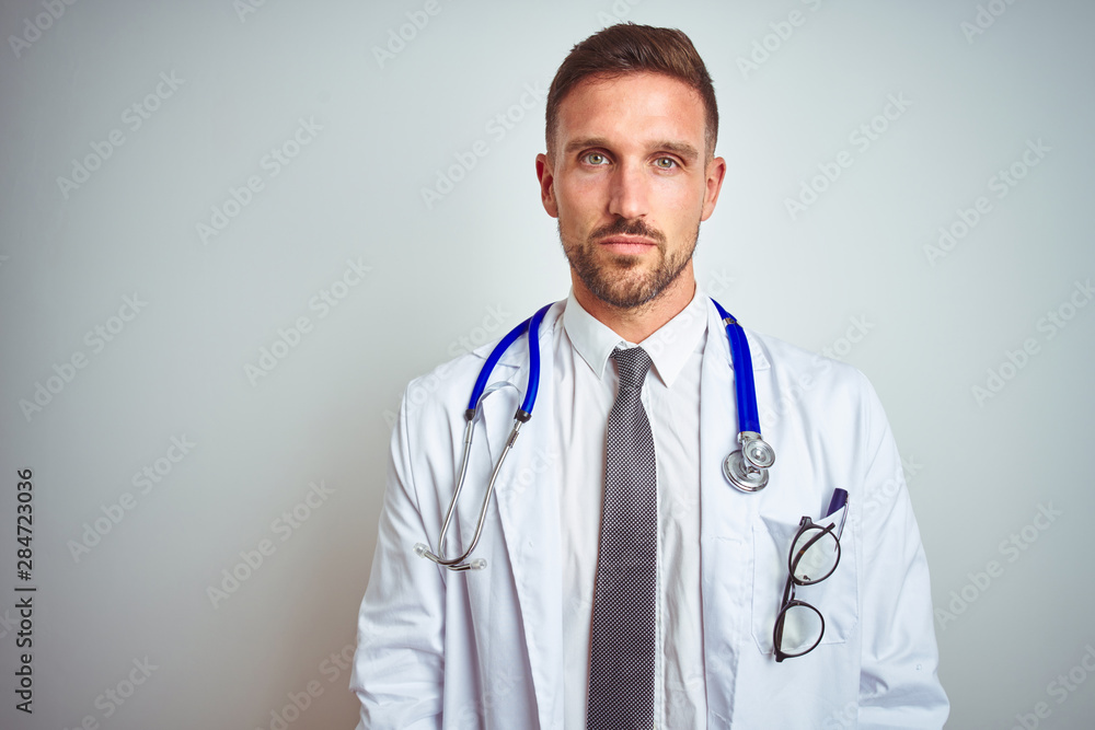 Young handsome doctor man wearing white professional coat over isolated background with serious expression on face. Simple and natural looking at the camera. Young handsome doctor man wearing white professional coat over isolated background with serious expression on face. Simple and natural looking at the camera.