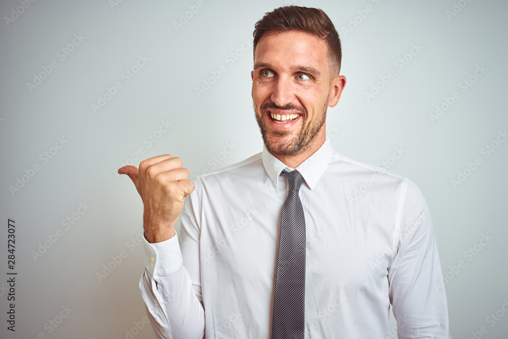Young handsome business man wearing elegant white shirt over isolated background smiling with happy face looking and pointing to the side with thumb up. Young handsome business man wearing elegant white shirt over isolated background smiling with happy face looking and pointing to the side with thumb up.