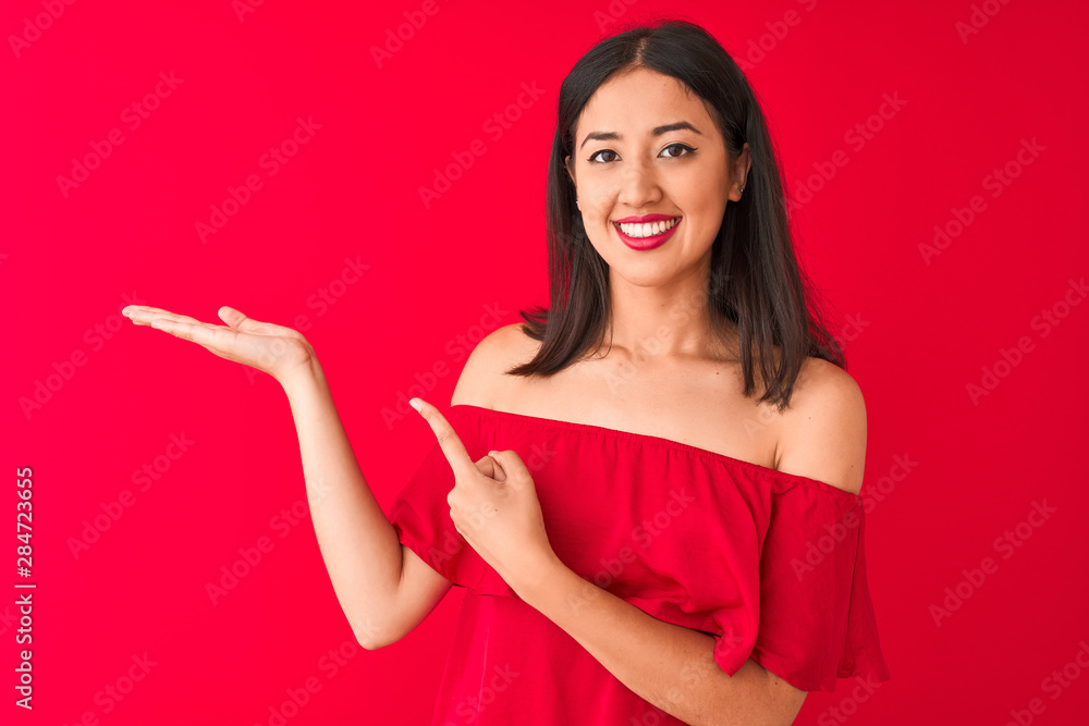 Young beautiful chinese woman wearing casual t-shirt standing over isolated red background amazed and smiling to the camera while presenting with hand and pointing with finger. Young beautiful chinese woman wearing casual t-shirt standing over isolated red background amazed and smiling to the camera while presenting with hand and pointing with finger.