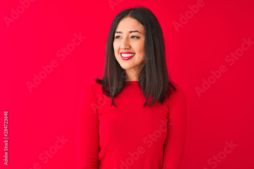 Young beautiful chinese woman wearing casual dress standing over isolated red background looking away to side with smile on face, natural expression. Laughing confident.