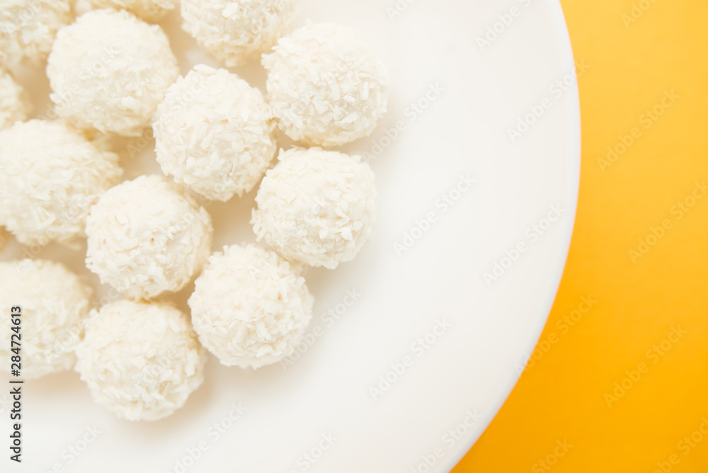 Chocolate covered coconut candies on white plate isolated on yellow background. Truffle candy set. Selective focus Chocolate covered coconut candies on white plate isolated on yellow background. Truffle candy set. Selective focus