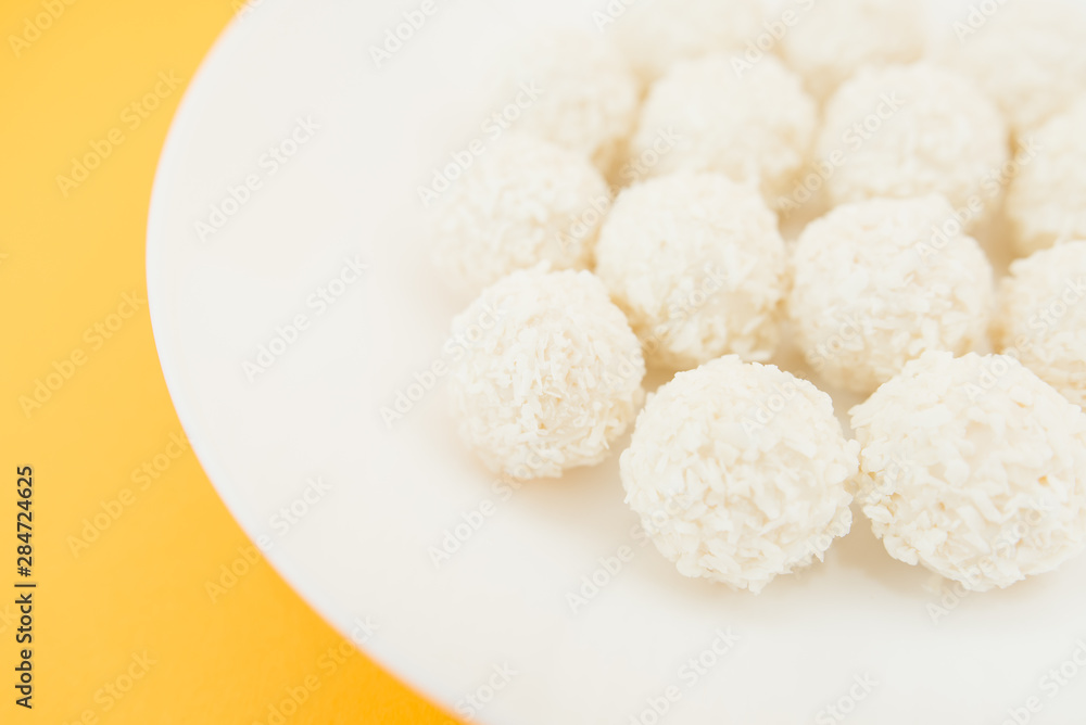 Chocolate covered coconut candies on white plate isolated on yellow background. Truffle candy set. Selective focus Chocolate covered coconut candies on white plate isolated on yellow background. Truffle candy set. Selective focus