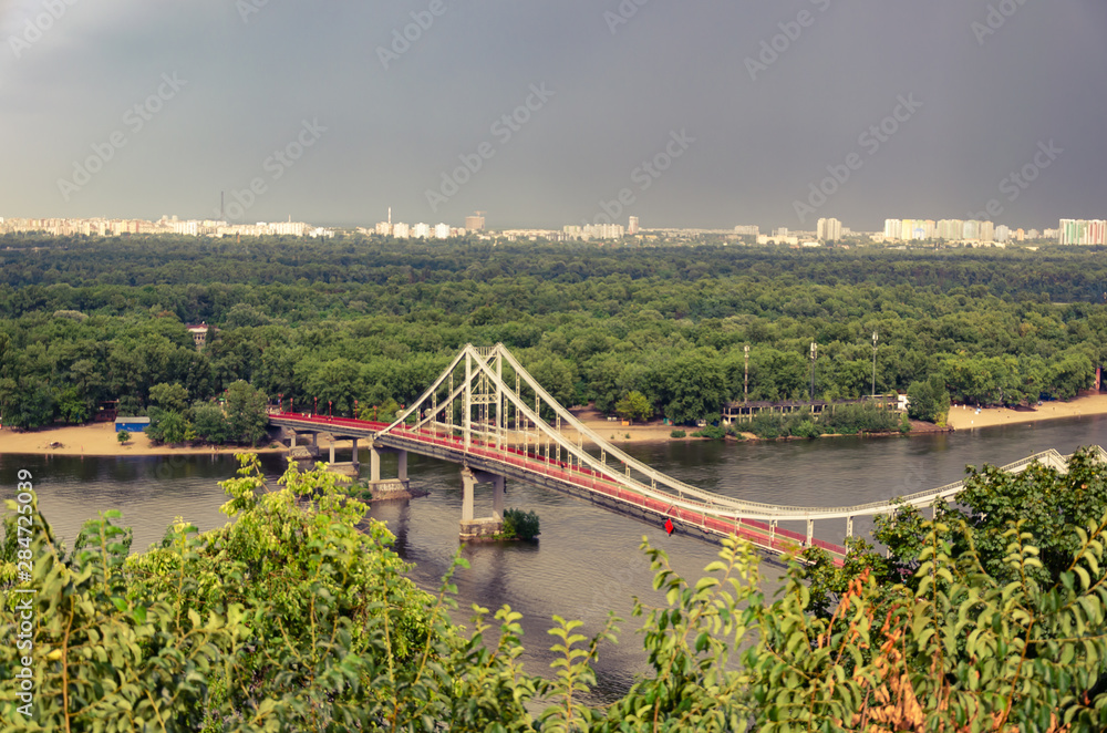 View of pedestrian Park bridge over the Dnipro river. Cloudy sky after rain. Summertime