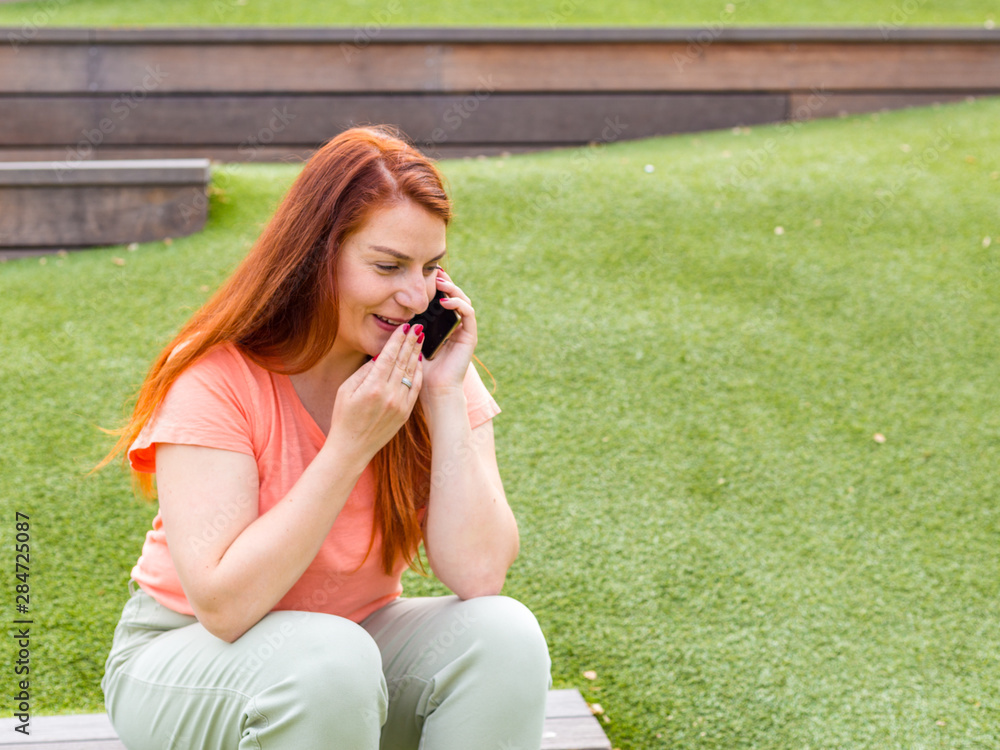 Smiling caucasian woman talking on mobile phone. Happy young ginger woman using smart phone outdoors, girl sitting on a bench in the park. Concept of generation Smiling caucasian woman talking on mobile phone. Happy young ginger woman using smart phone outdoors, girl sitting on a bench in the park. Concept of generation