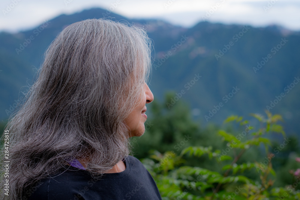 Graceful Indian ethnicity woman with gray hair standing in sports clothing against a scenic hilly backdrop.