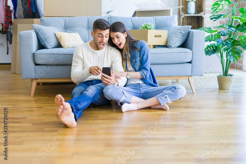 Young couple sitting on the floor of new home using smartphone and smiling happy for moving to a new home