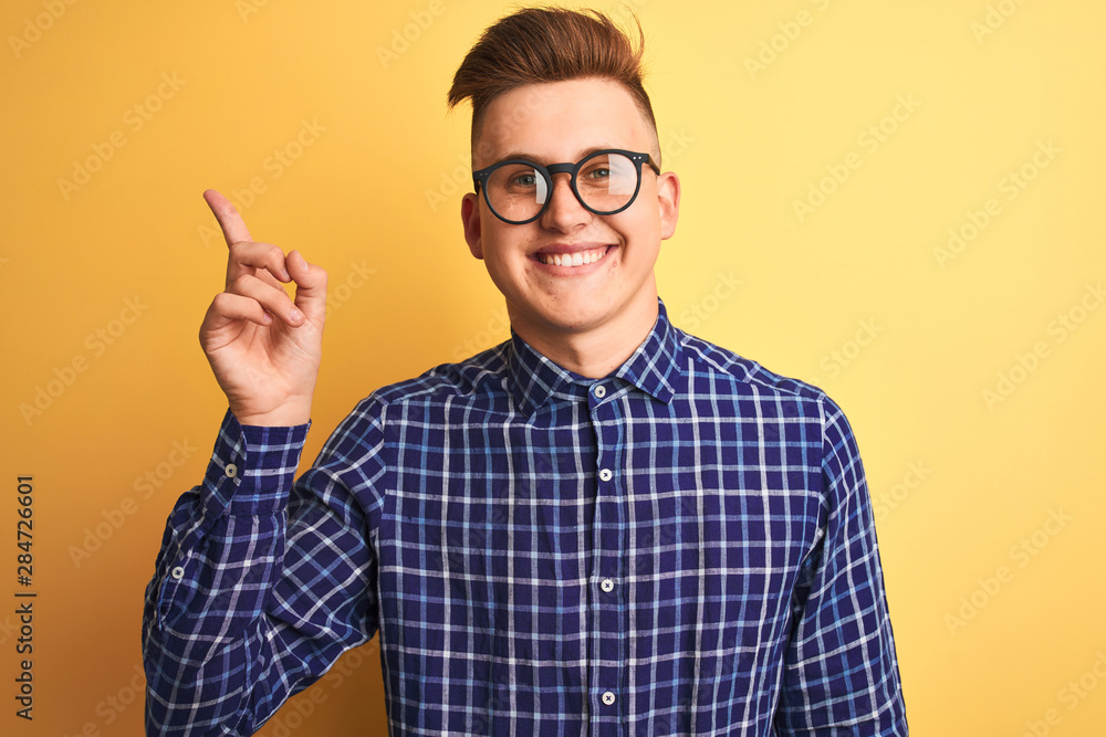 Young handsome man wearing casual shirt and glasses over isolated yellow background with a big smile on face, pointing with hand and finger to the side looking at the camera. Young handsome man wearing casual shirt and glasses over isolated yellow background with a big smile on face, pointing with hand and finger to the side looking at the camera.