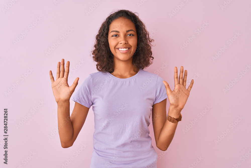 Young brazilian woman wearing t-shirt standing over isolated pink background showing and pointing up with fingers number ten while smiling confident and happy. Young brazilian woman wearing t-shirt standing over isolated pink background showing and pointing up with fingers number ten while smiling confident and happy.
