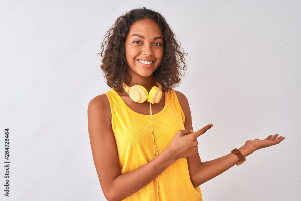 Young brazilian woman wearing yellow headphones over isolated white background amazed and smiling to the camera while presenting with hand and pointing with finger. Young brazilian woman wearing yellow headphones over isolated white background amazed and smiling to the camera while presenting with hand and pointing with finger.