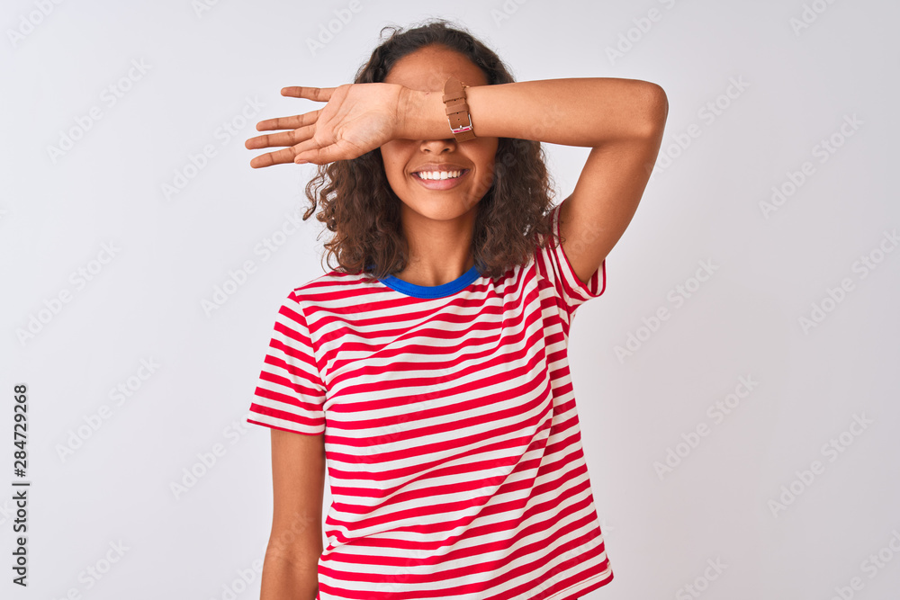 Young brazilian woman wearing red striped t-shirt standing over isolated white background covering eyes with arm smiling cheerful and funny. Blind concept. Young brazilian woman wearing red striped t-shirt standing over isolated white background covering eyes with arm smiling cheerful and funny. Blind concept.