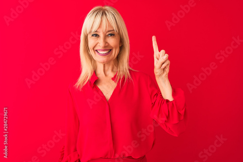 Middle age woman wearing elegant shirt standing over isolated red background showing and pointing up with finger number one while smiling confident and happy.