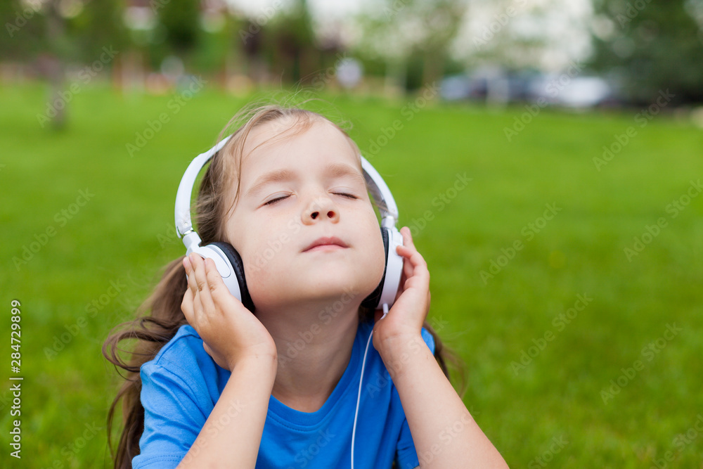 little cute girl sitting in the Park on the grass on a Sunny summer day and listening to music in white headphones. Lifestyle, happy summertime, summer outdoor portrait little cute girl sitting in the Park on the grass on a Sunny summer day and listening to music in white headphones. Lifestyle, happy summertime, summer outdoor portrait