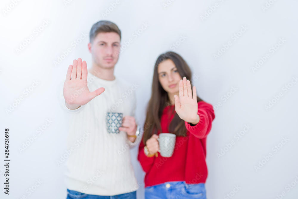 Beautiful young couple over white isolated background drinking a cup of coffee with open hand doing stop sign with serious and confident expression, defense gesture