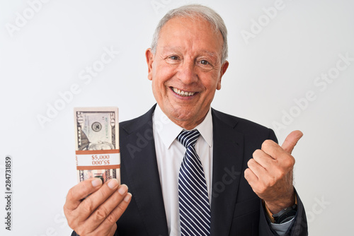 Senior grey-haired businessman wearing suit holding dollars over isolated white background pointing and showing with thumb up to the side with happy face smiling