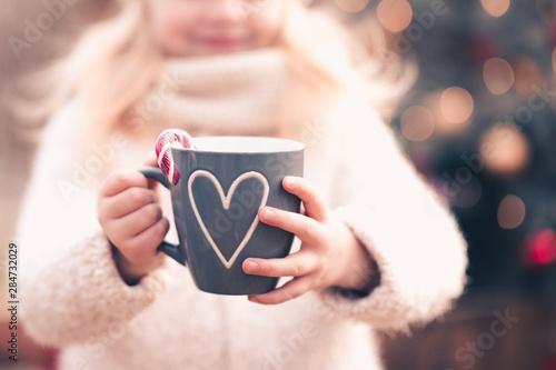 Child girl holding Christmas cup of tea with candy cane outdoors over lights at background close up. Winter holidays.