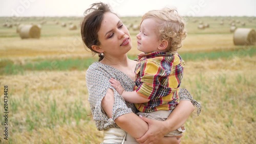 Beautiful young woman with fair-haired little son in her arms in the middle of a wheat field with bales in summer evening. Mom kisses her son