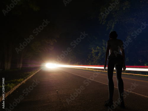 A young woman or girl all alone standing beside a road at night after dark with a long exposure background of car lights streaking by in a forest deep in the secluded hills and woods of Tennessee.