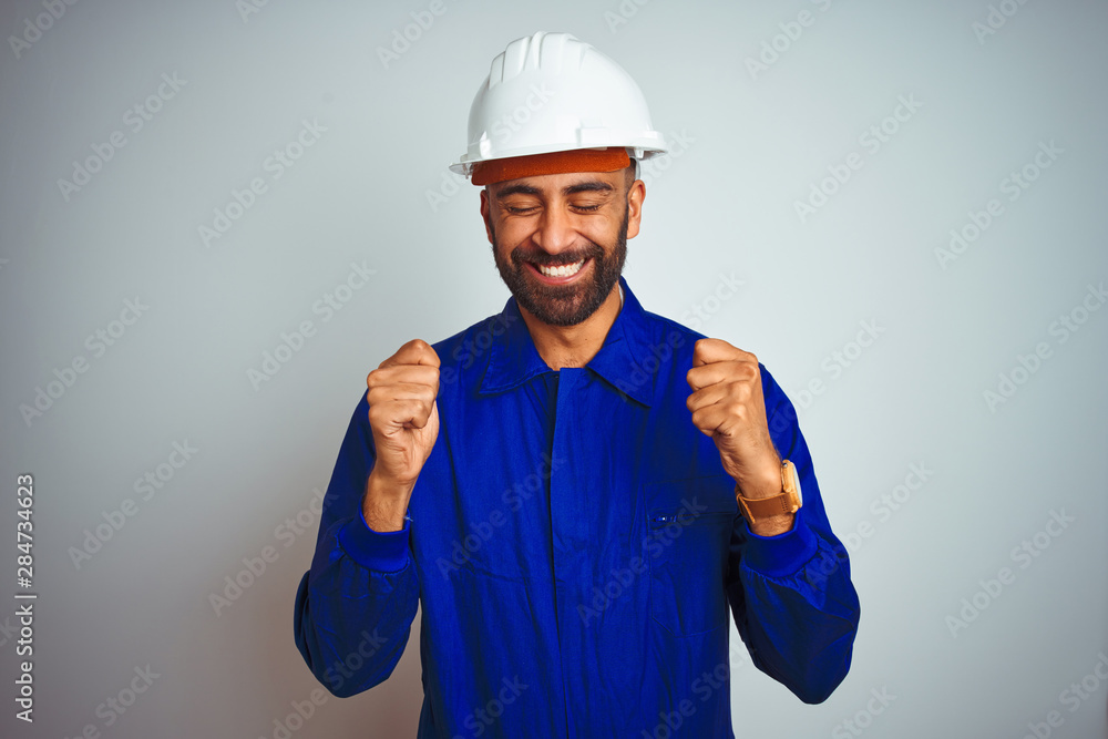 Handsome indian worker man wearing uniform and helmet over isolated white background excited for success with arms raised and eyes closed celebrating victory smiling. Winner concept.