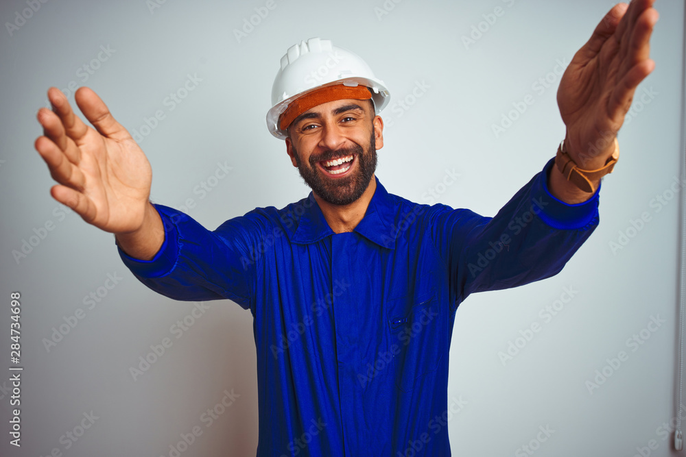 Handsome indian worker man wearing uniform and helmet over isolated white background looking at the camera smiling with open arms for hug. Cheerful expression embracing happiness.