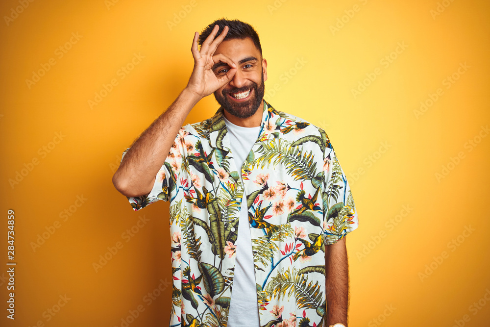 Fototapeta premium Young indian man on vacation wearing summer floral shirt over isolated yellow background doing ok gesture with hand smiling, eye looking through fingers with happy face.