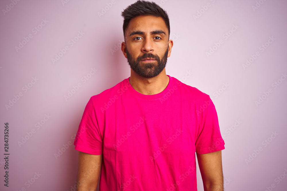 Young indian man wearing t-shirt standing over isolated pink background Relaxed with serious expression on face. Simple and natural looking at the camera. Young indian man wearing t-shirt standing over isolated pink background Relaxed with serious expression on face. Simple and natural looking at the camera.