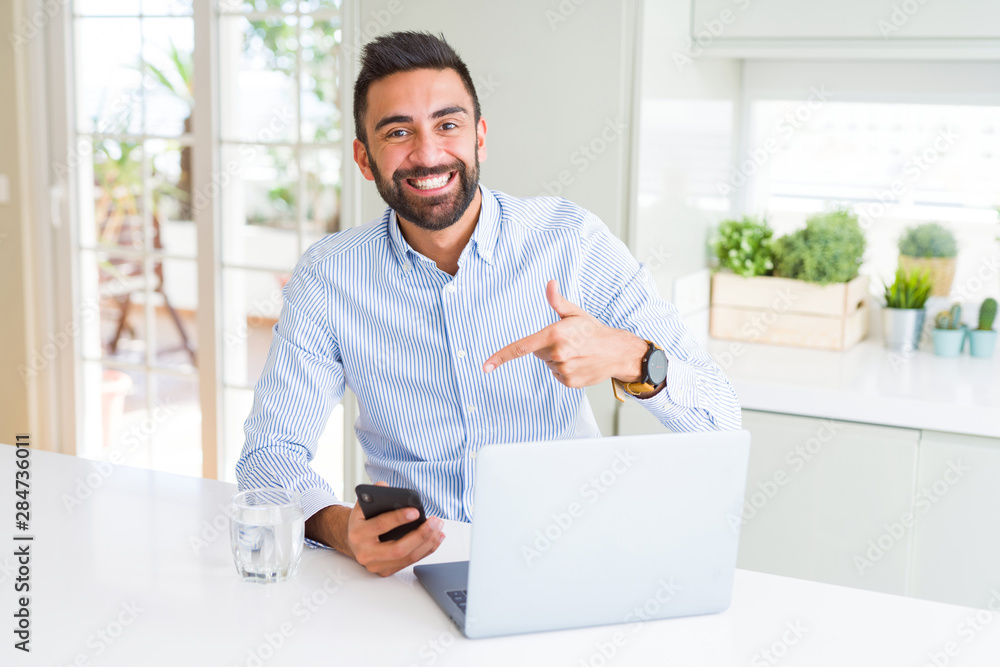 Handsome hispanic business man using laptop and smartphone very happy pointing with hand and finger