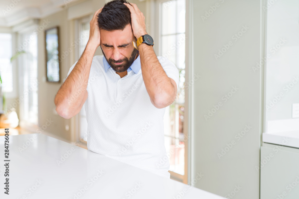 Handsome hispanic man casual white t-shirt at home suffering from headache desperate and stressed because pain and migraine. Hands on head. Handsome hispanic man casual white t-shirt at home suffering from headache desperate and stressed because pain and migraine. Hands on head.