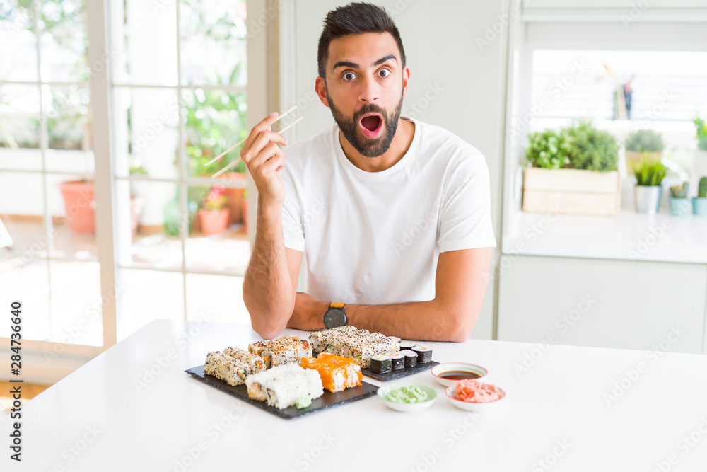 Handsome hispanic man eating asian sushi using chopsticks scared in shock with a surprise face, afraid and excited with fear expression