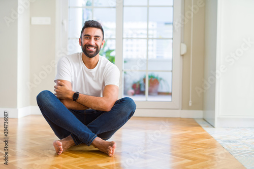 Handsome hispanic man wearing casual t-shirt sitting on the floor at home happy face smiling with crossed arms looking at the camera. Positive person.