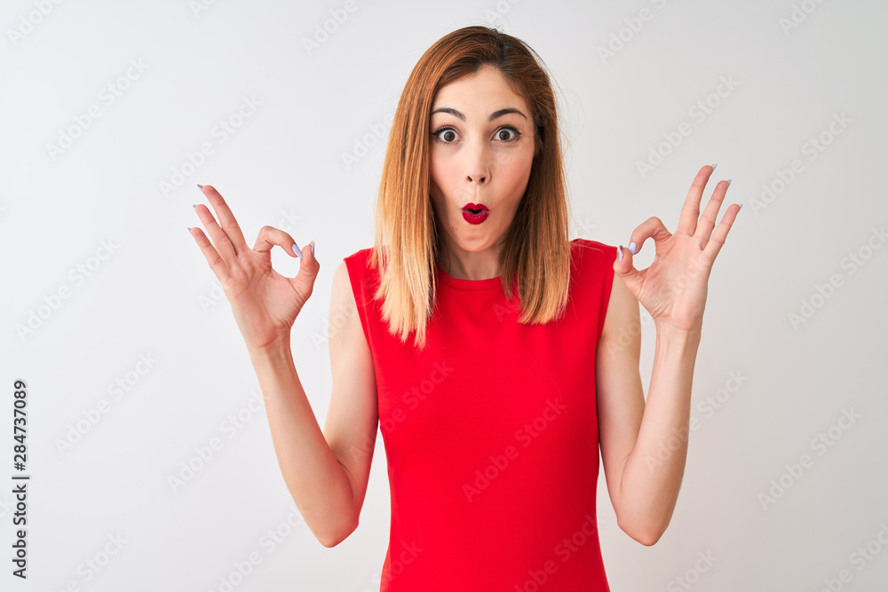 Redhead businesswoman wearing elegant red dress standing over isolated white background looking surprised and shocked doing ok approval symbol with fingers. Crazy expression Redhead businesswoman wearing elegant red dress standing over isolated white background looking surprised and shocked doing ok approval symbol with fingers. Crazy expression