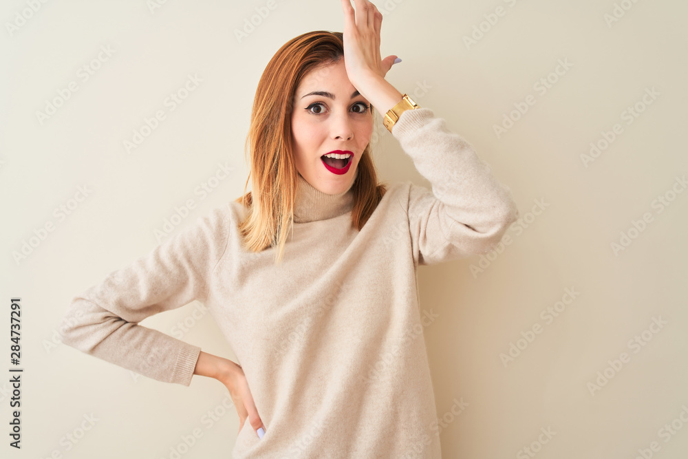 Fototapeta premium Redhead woman wearing elegant turtleneck sweater standing over isolated white background surprised with hand on head for mistake, remember error. Forgot, bad memory concept.