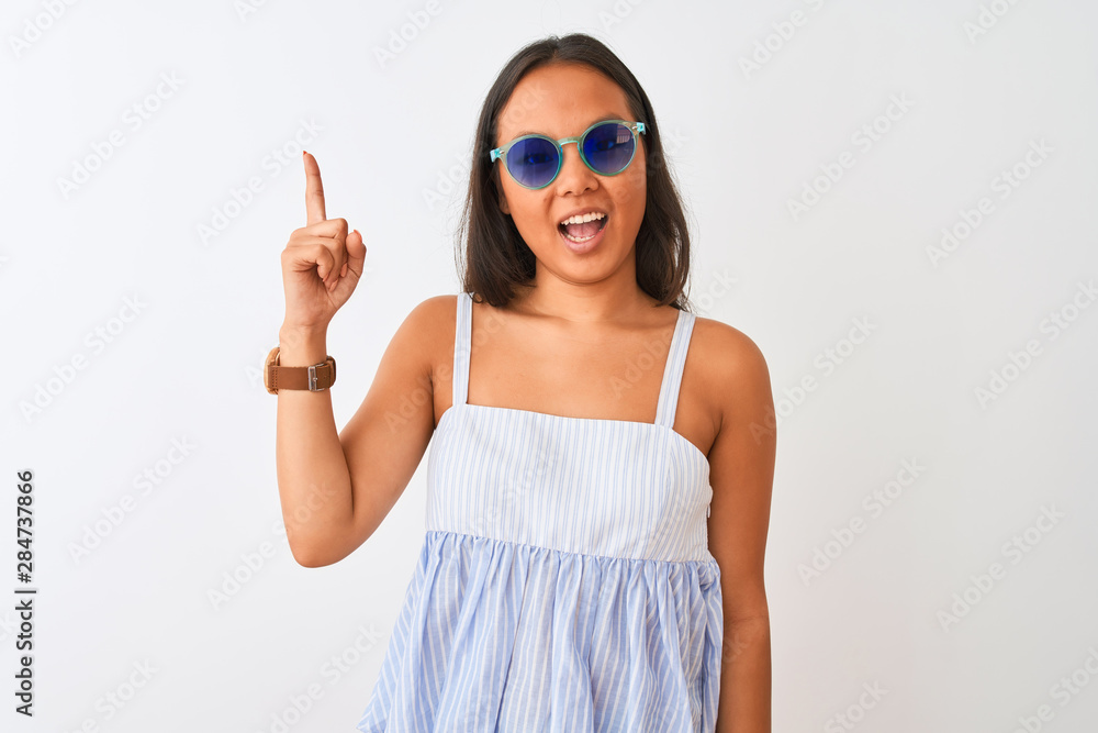 Young chinese woman wearing striped dress and sunglasses over isolated white background pointing finger up with successful idea. Exited and happy. Number one. Young chinese woman wearing striped dress and sunglasses over isolated white background pointing finger up with successful idea. Exited and happy. Number one.