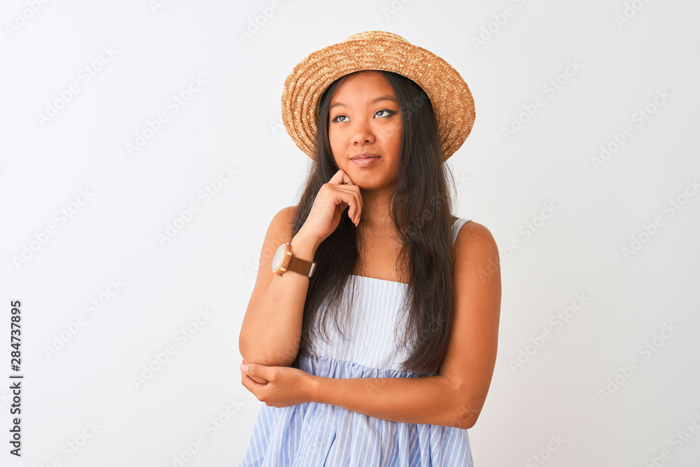 Young chinese woman wearing striped dress and hat standing over isolated white background with hand on chin thinking about question, pensive expression. Smiling with thoughtful face. Doubt concept. Young chinese woman wearing striped dress and hat standing over isolated white background with hand on chin thinking about question, pensive expression. Smiling with thoughtful face. Doubt concept.