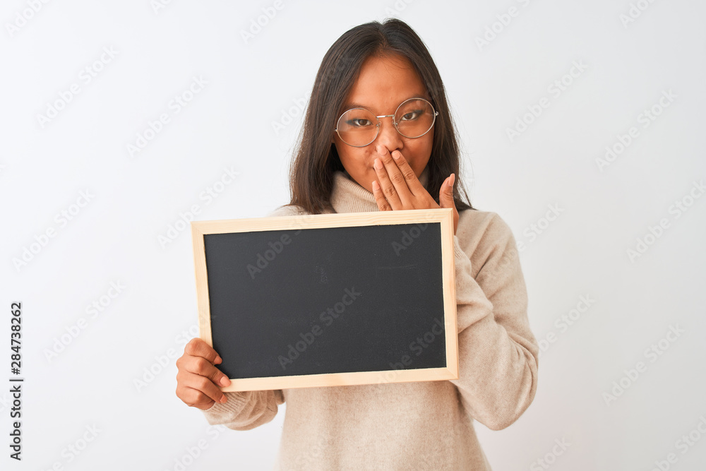 Young chinese woman wearing glasses holding blackboard over isolated white background cover mouth with hand shocked with shame for mistake, expression of fear, scared in silence, secret concept
