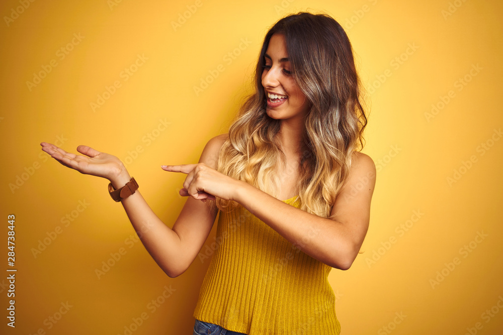 Young beautiful woman wearing t-shirt over yellow isolated background amazed and smiling to the camera while presenting with hand and pointing with finger. Young beautiful woman wearing t-shirt over yellow isolated background amazed and smiling to the camera while presenting with hand and pointing with finger.