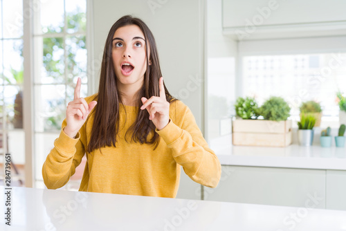 Beautiful young woman wearing yellow sweater amazed and surprised looking up and pointing with fingers and raised arms.