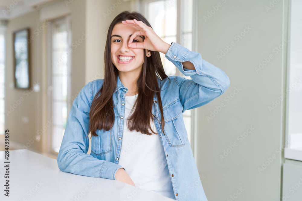 Beautiful young woman sitting on white table at home doing ok gesture with hand smiling, eye looking through fingers with happy face. Beautiful young woman sitting on white table at home doing ok gesture with hand smiling, eye looking through fingers with happy face.