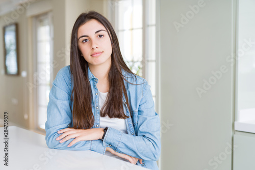 Beautiful young woman sitting on white table at home with serious expression on face. Simple and natural looking at the camera.