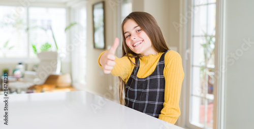 Beautiful young girl kid sitting on the table doing happy thumbs up gesture with hand. Approving expression looking at the camera showing success.