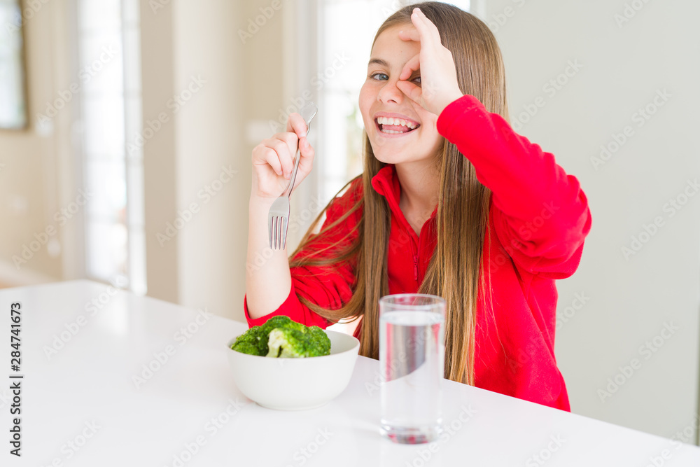 Beautiful young girl kid eating fresh broccoli and drinking water with happy face smiling doing ok sign with hand on eye looking through fingers Beautiful young girl kid eating fresh broccoli and drinking water with happy face smiling doing ok sign with hand on eye looking through fingers