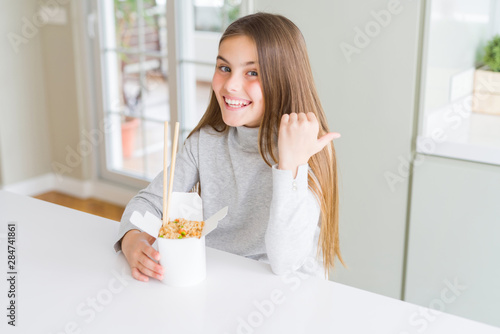 Beautiful young girl kid eating asian rice in delivery box pointing and showing with thumb up to the side with happy face smiling