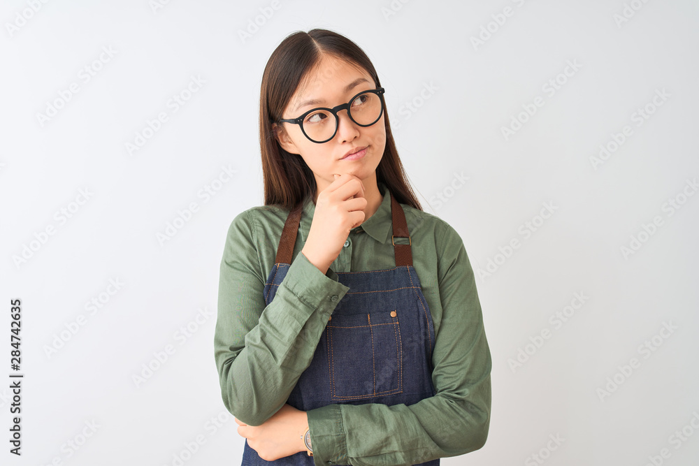 Young chinese shopkeeper woman wearing apron and glasses over isolated white background with hand on chin thinking about question, pensive expression. Smiling with thoughtful face. Doubt concept. Young chinese shopkeeper woman wearing apron and glasses over isolated white background with hand on chin thinking about question, pensive expression. Smiling with thoughtful face. Doubt concept.