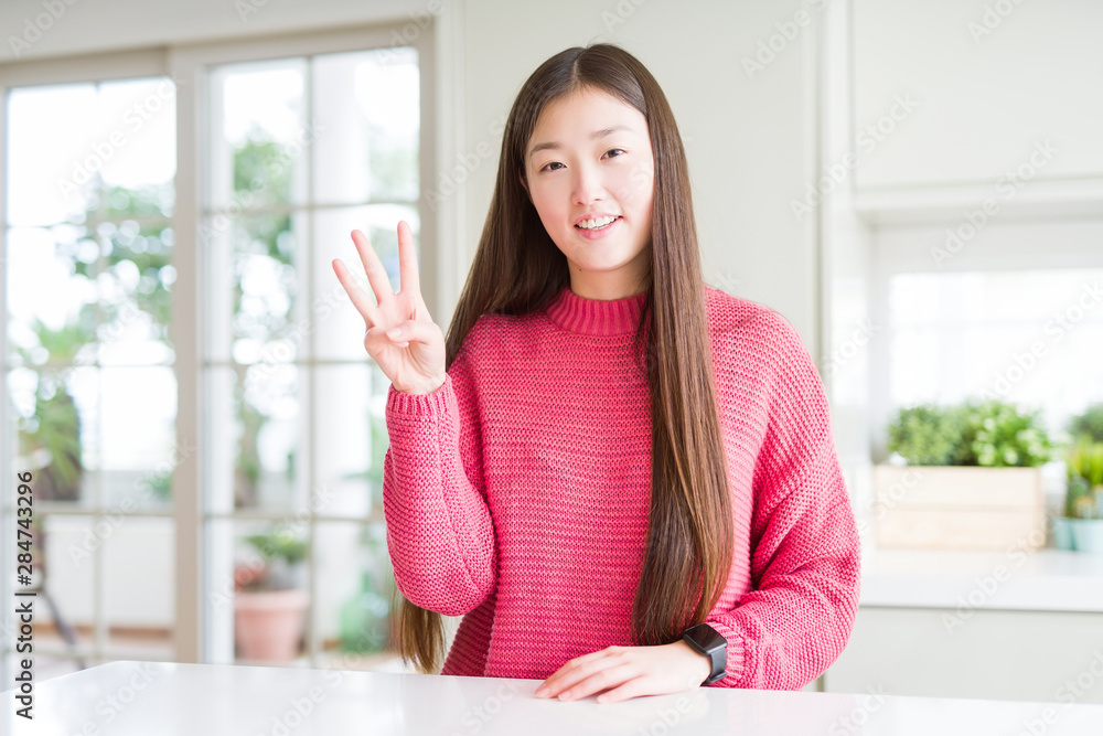 Beautiful Asian woman wearing pink sweater on white table showing and pointing up with fingers number three while smiling confident and happy.