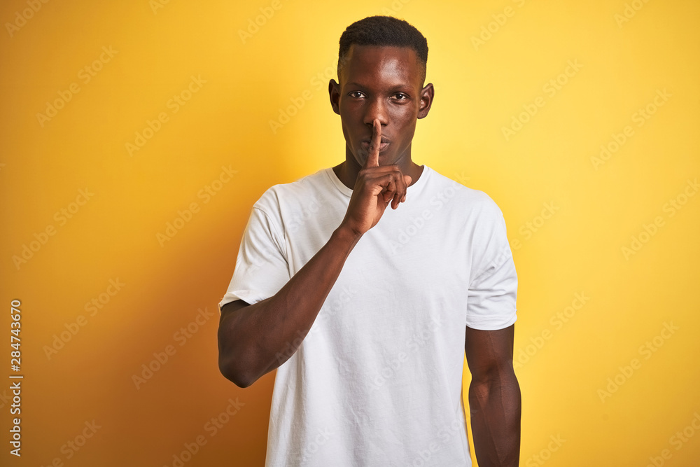 Young african american man wearing white t-shirt standing over isolated yellow background asking to be quiet with finger on lips. Silence and secret concept. Young african american man wearing white t-shirt standing over isolated yellow background asking to be quiet with finger on lips. Silence and secret concept.