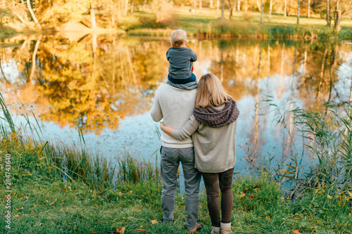 Lifestyle family from behind with their child on fathers shoulders standing at lake shore at sunset